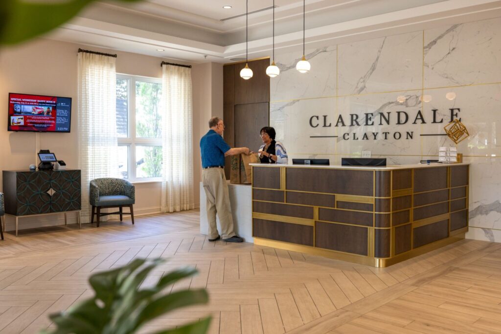 The main lobby of Clarendale Clayton in MO, a luxury retirment community. There is a receptionist greeting a senior citizen.