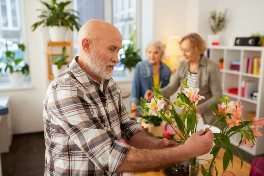 senior man arranging flower vase with two senior women in the background
